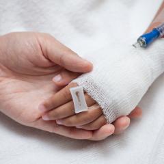 child's bandaged hand with drip point inserted held by adult hand on white sheet background